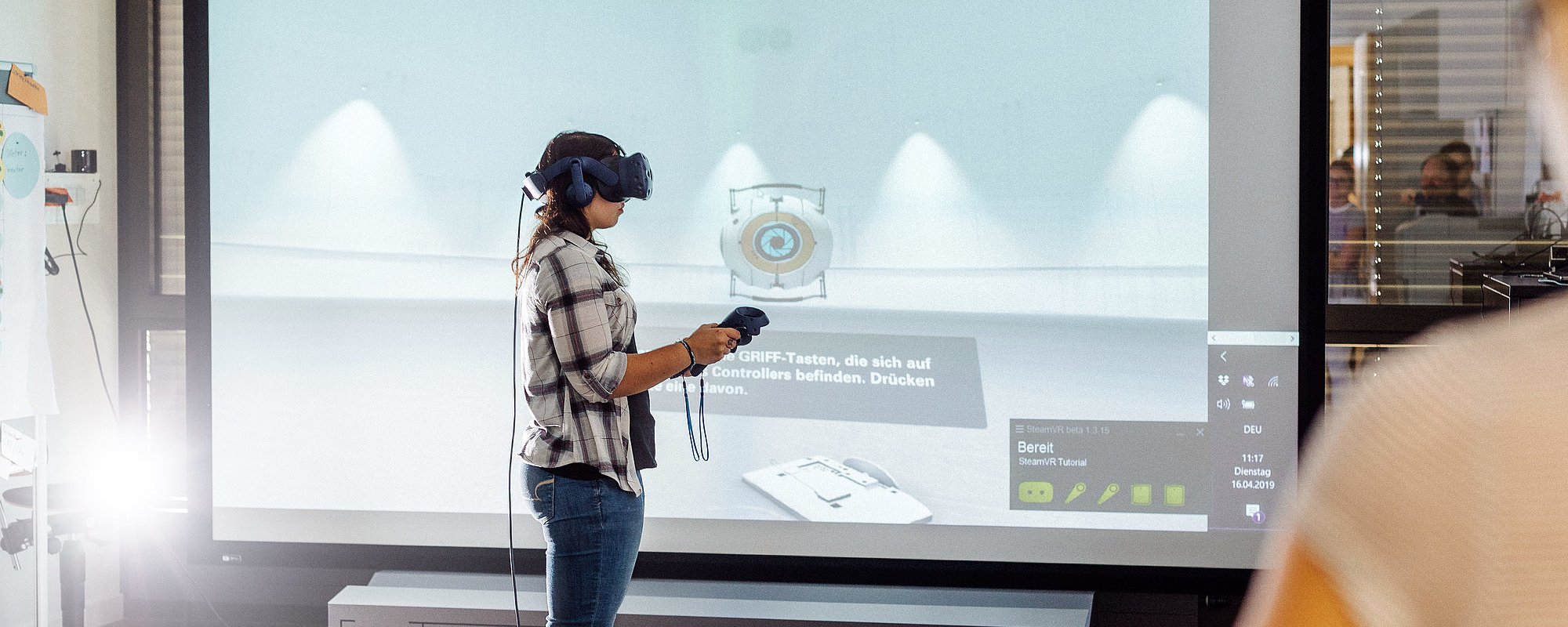 Person trägt VR-Brille und hält Joysticks in beiden Händen. Im Hintergrund ist ein großer Bildschirm zu sehen.