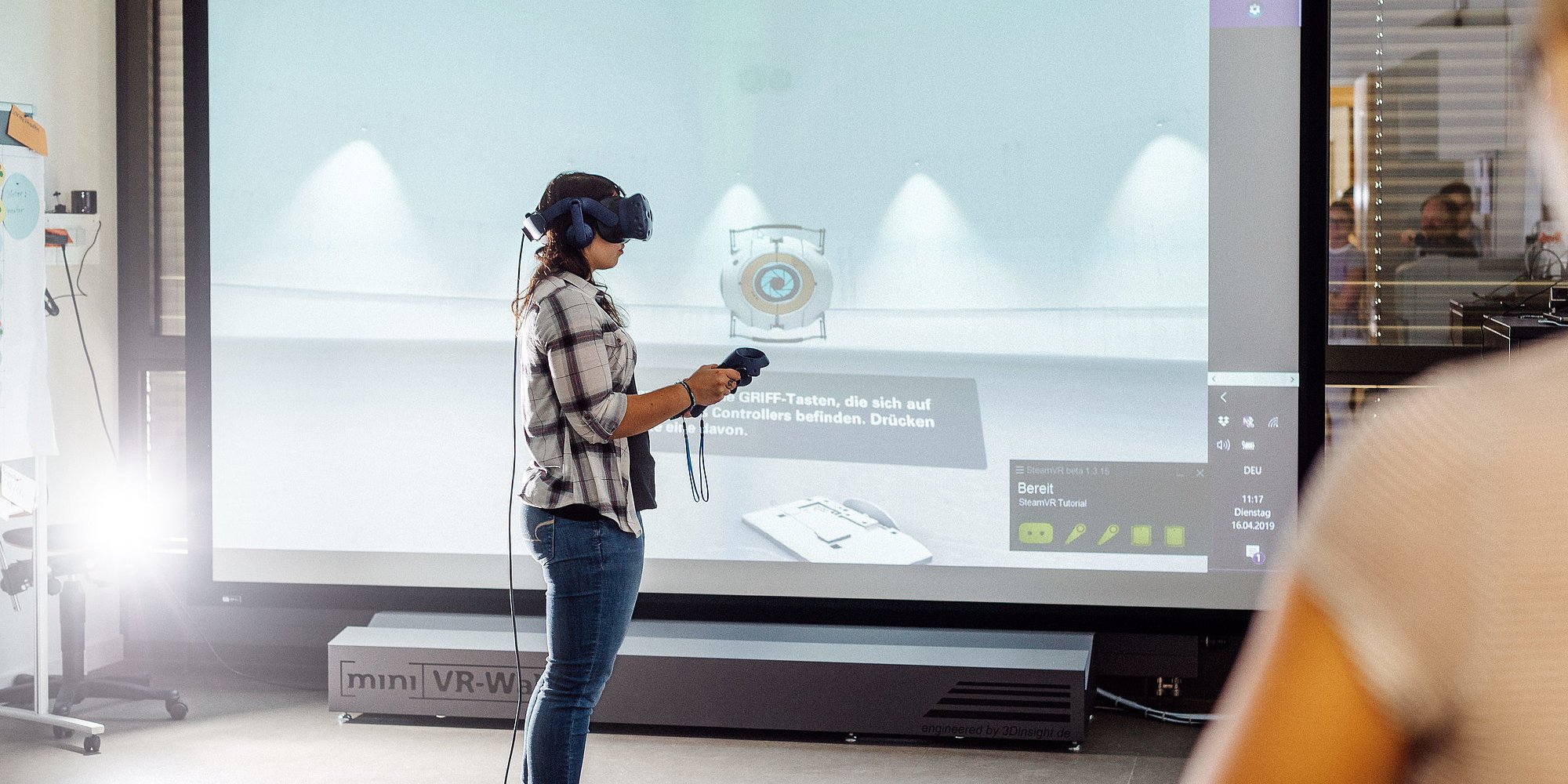 Person trägt VR-Brille und hält Joysticks in beiden Händen. Im Hintergrund ist ein großer Bildschirm zu sehen.