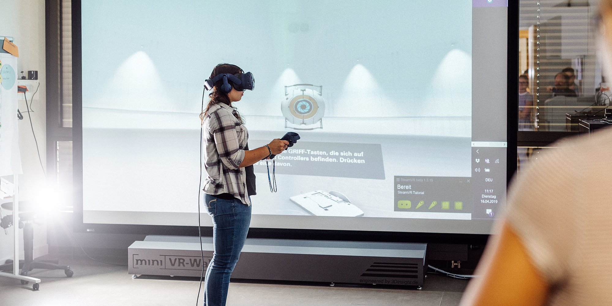 Person trägt VR-Brille und hält Joysticks in beiden Händen. Im Hintergrund ist ein großer Bildschirm zu sehen.